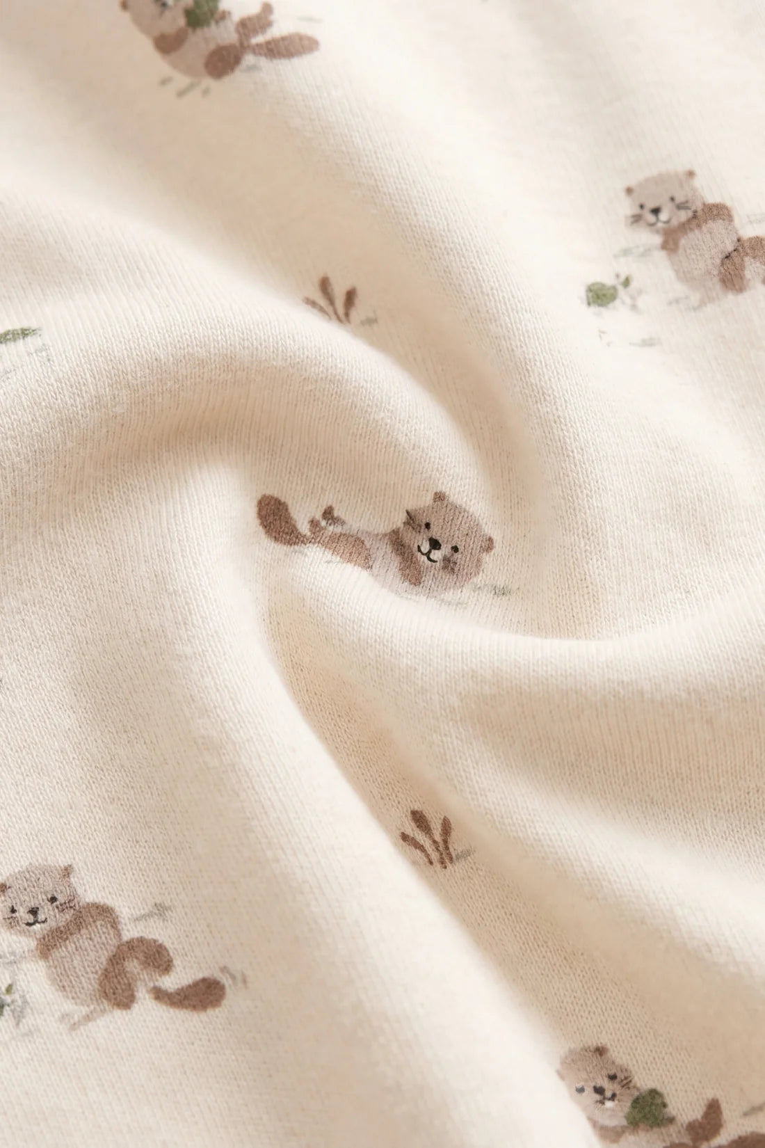 Close-up of a soft cream-colored fabric with a printed pattern of small brown bears and tiny green leaves, shown with gentle folds.