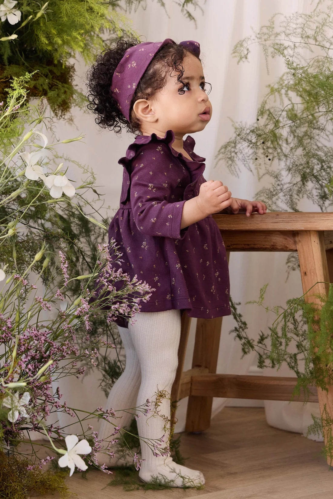 A toddler girl wearing a purple long-sleeve dress with a small floral pattern, a matching purple headband, and white tights, standing next to a wooden bench in a decorated indoor setting with greenery and flowers.
