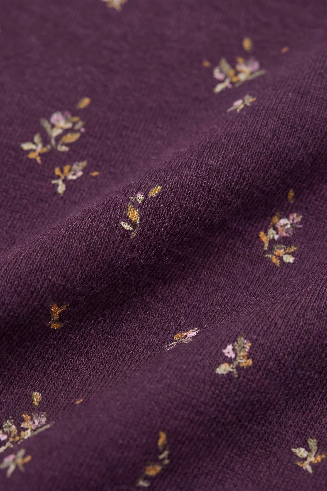 A close-up of a knitted fabric in deep plum purple, featuring a delicate, scattered floral pattern in muted yellow, pink, and white.