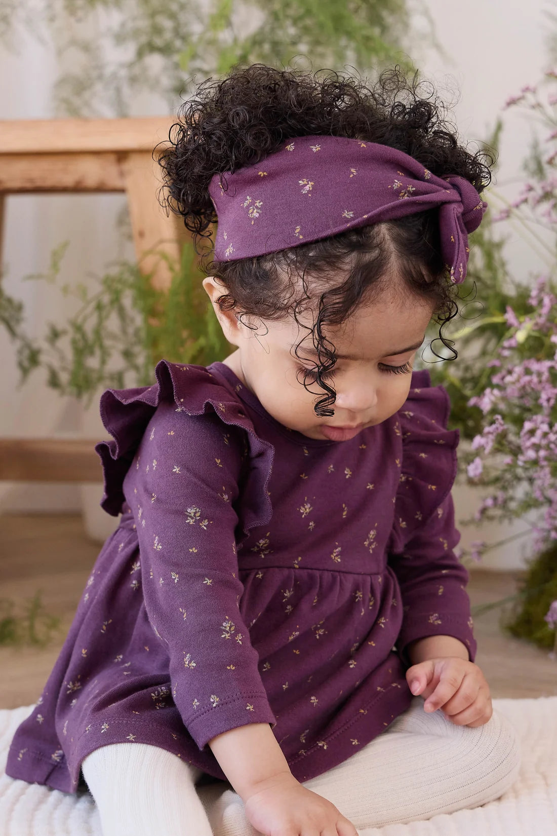 A toddler with curly dark hair wearing a purple long-sleeve dress with small gold floral prints, a matching purple headband, and white tights, sitting in a softly lit indoor setting with plants and a wooden bench in the background.