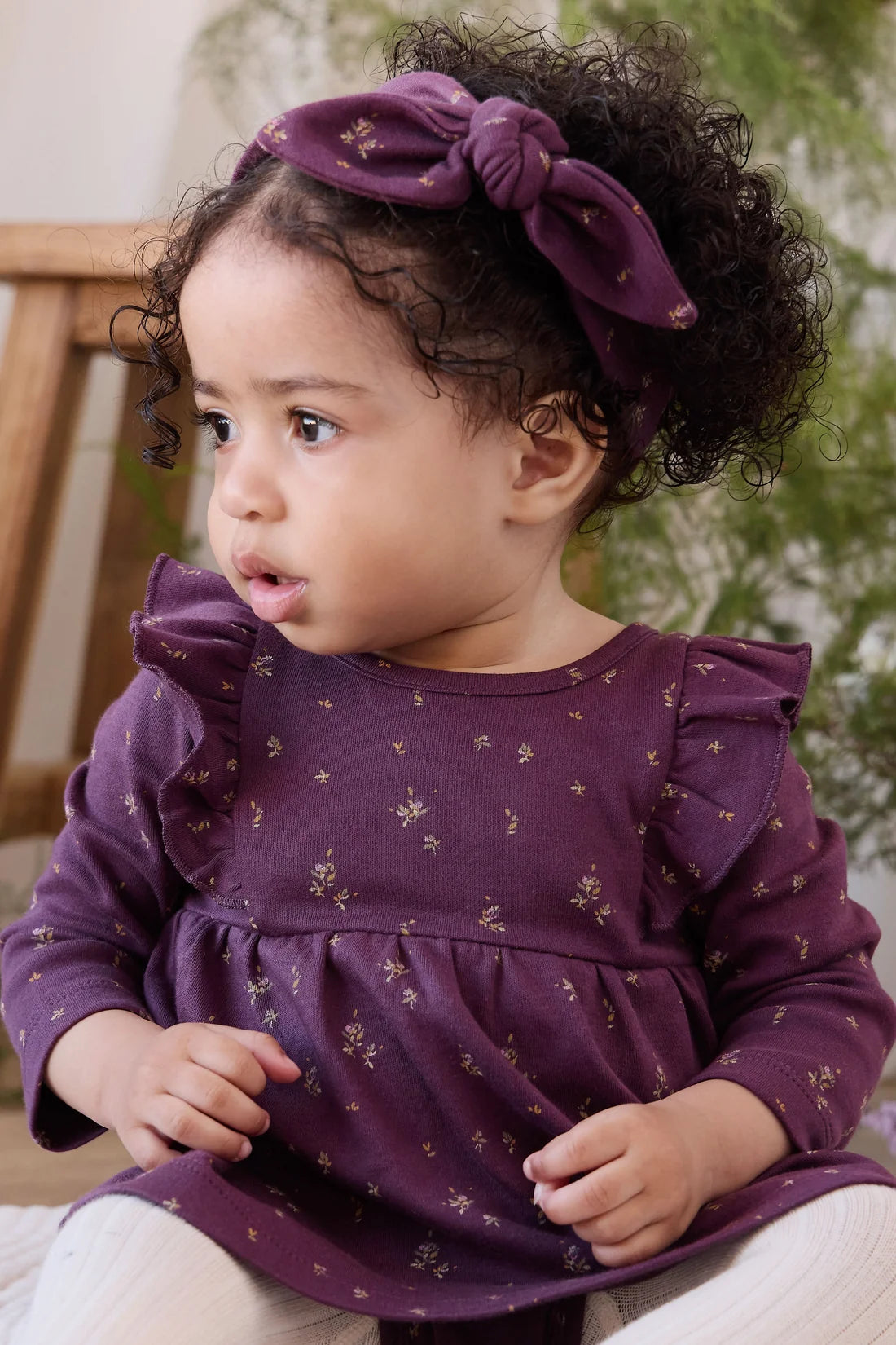 A toddler with curly dark hair wearing a purple fabric headband with a large bow and a matching purple long-sleeve top with small gold floral prints, seated indoors.