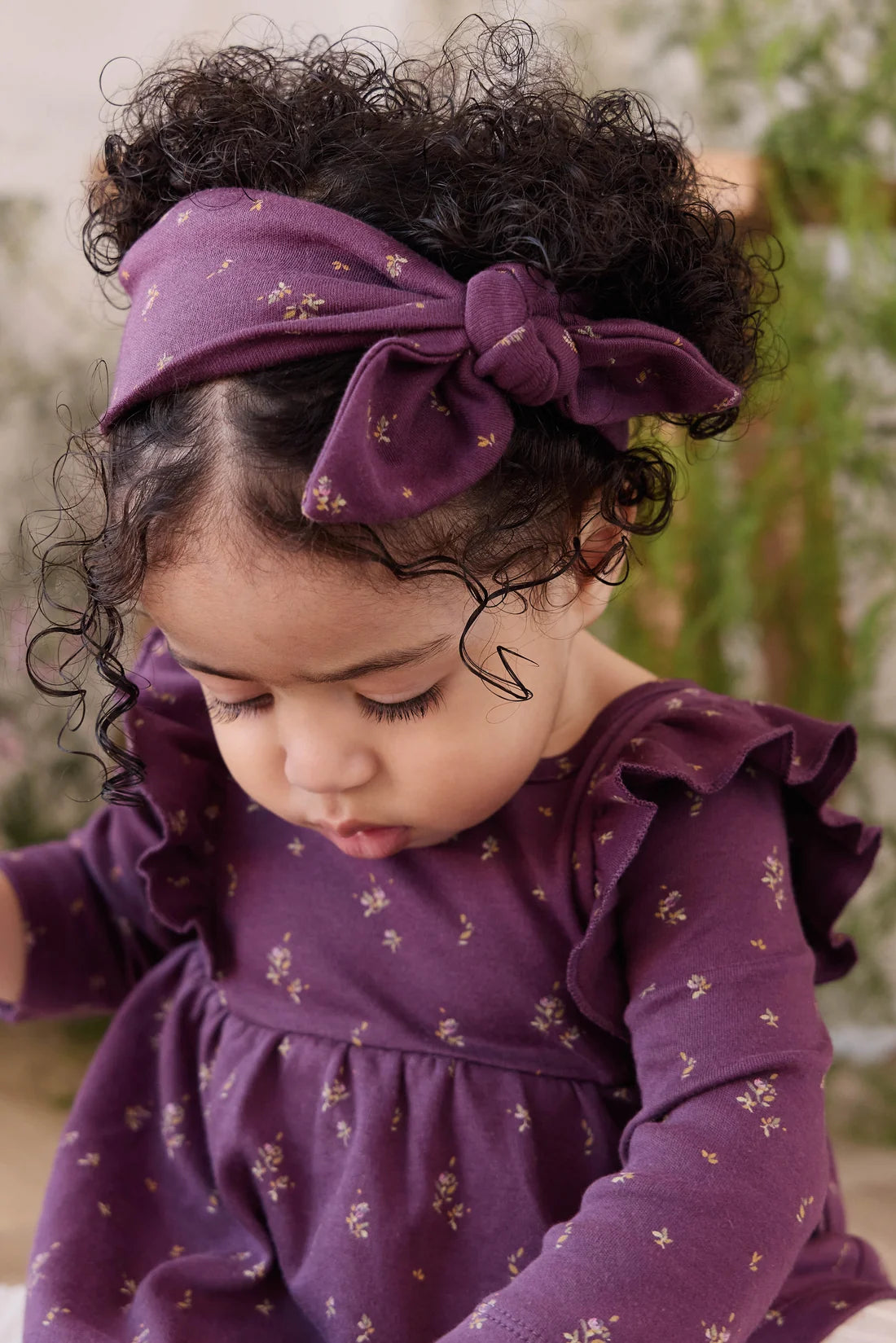 A toddler with curly dark hair wearing a purple fabric headband with a large bow and a matching purple long-sleeve dress with a small gold floral print, looking down in a softly blurred outdoor setting.