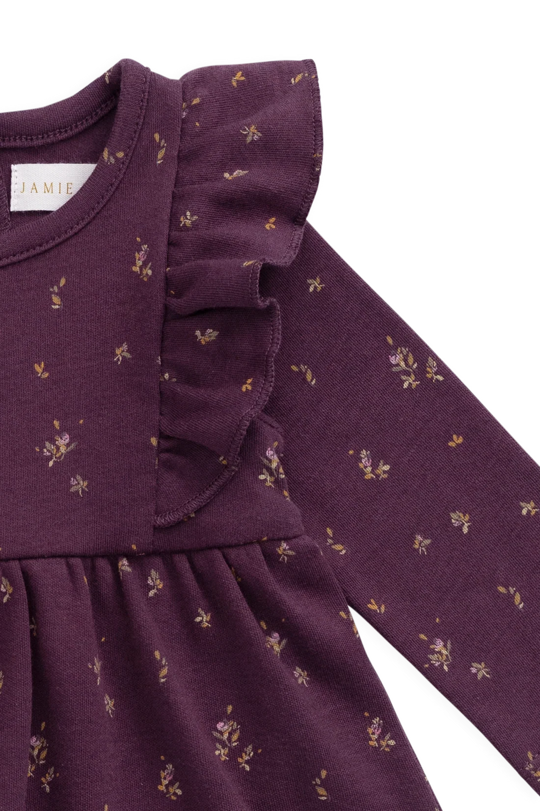 Close-up of a long-sleeve children's dress in deep plum purple with a small scattered floral print, featuring a ruffled neckline detail and a visible 'JAMIE' brand label.