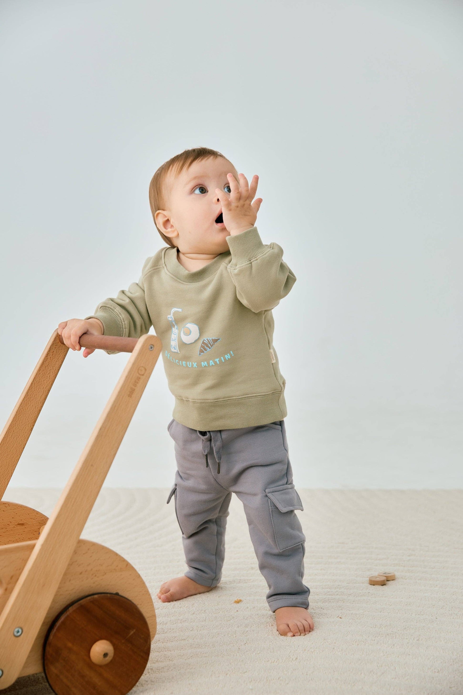 Baby in a green sweater and gray pants standing next to a wooden toy on a light-colored floor.