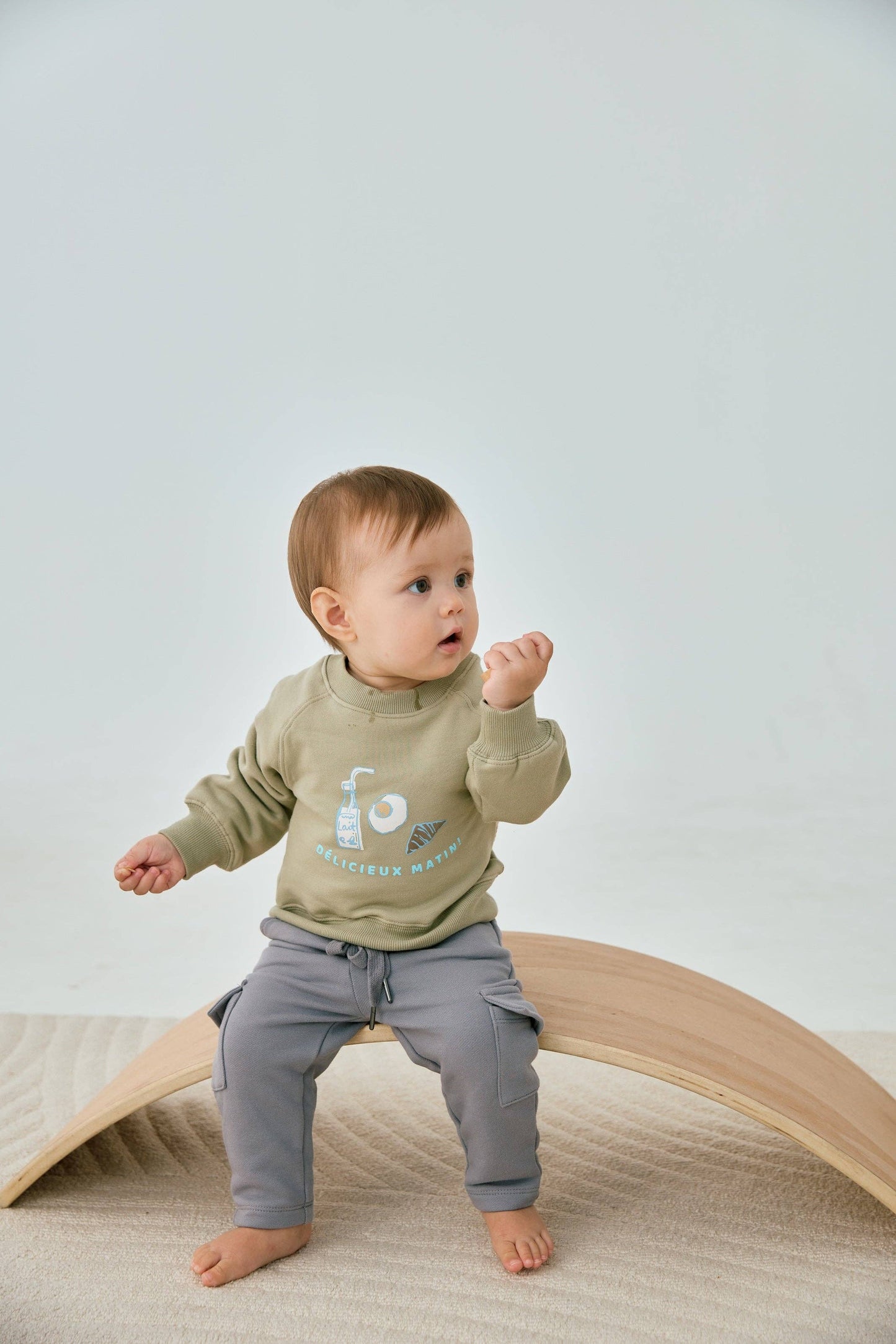 Baby wearing a green sweatshirt and gray pants standing on a wooden arch against a light background