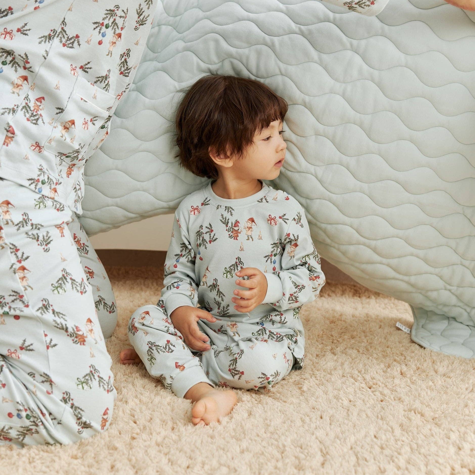 Child in floral pajamas sitting inside a homemade cardboard box fort with adult nearby.
