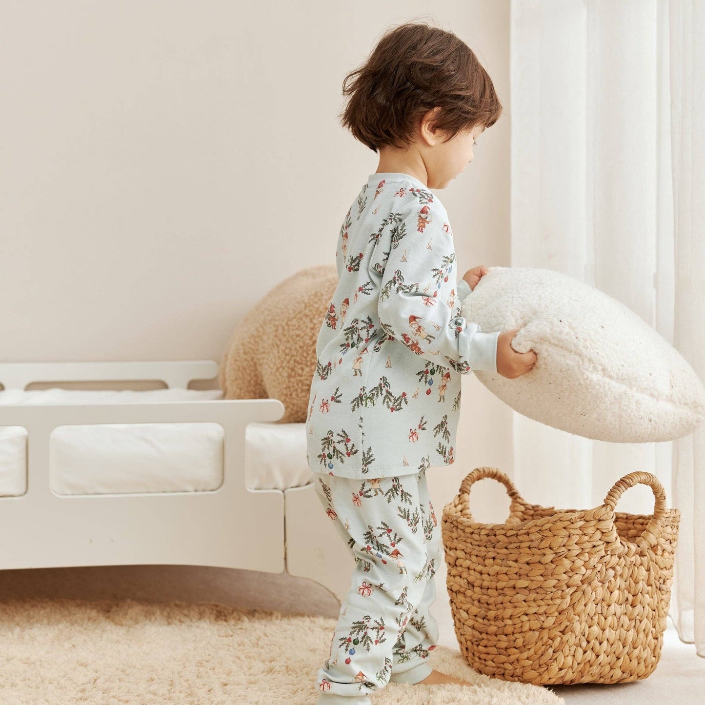 Child in floral pajamas holding a pillow in a room with a woven basket and white curtain.