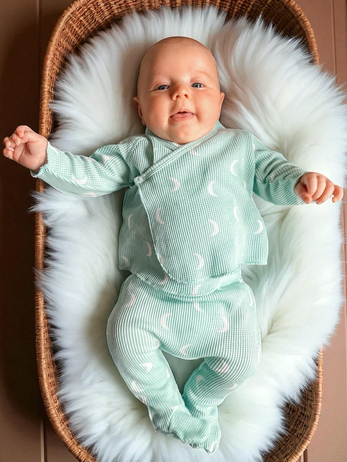 Baby in a mint green outfit lying on a fluffy white blanket inside a wooden basket.