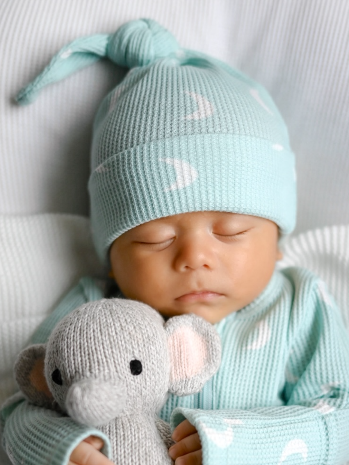 Baby wearing a light blue knit hat and matching outfit, holding a gray knitted elephant toy.