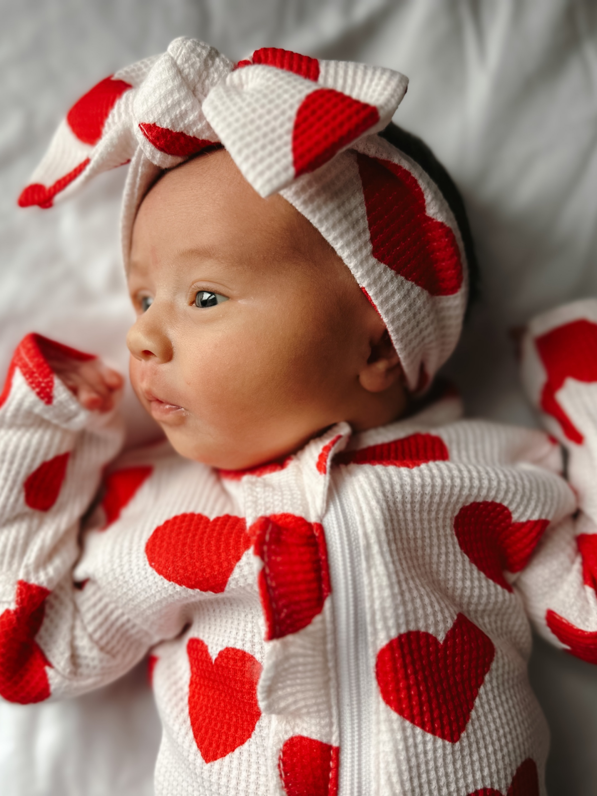 Baby wearing a white outfit with red hearts and a matching headband on a light background