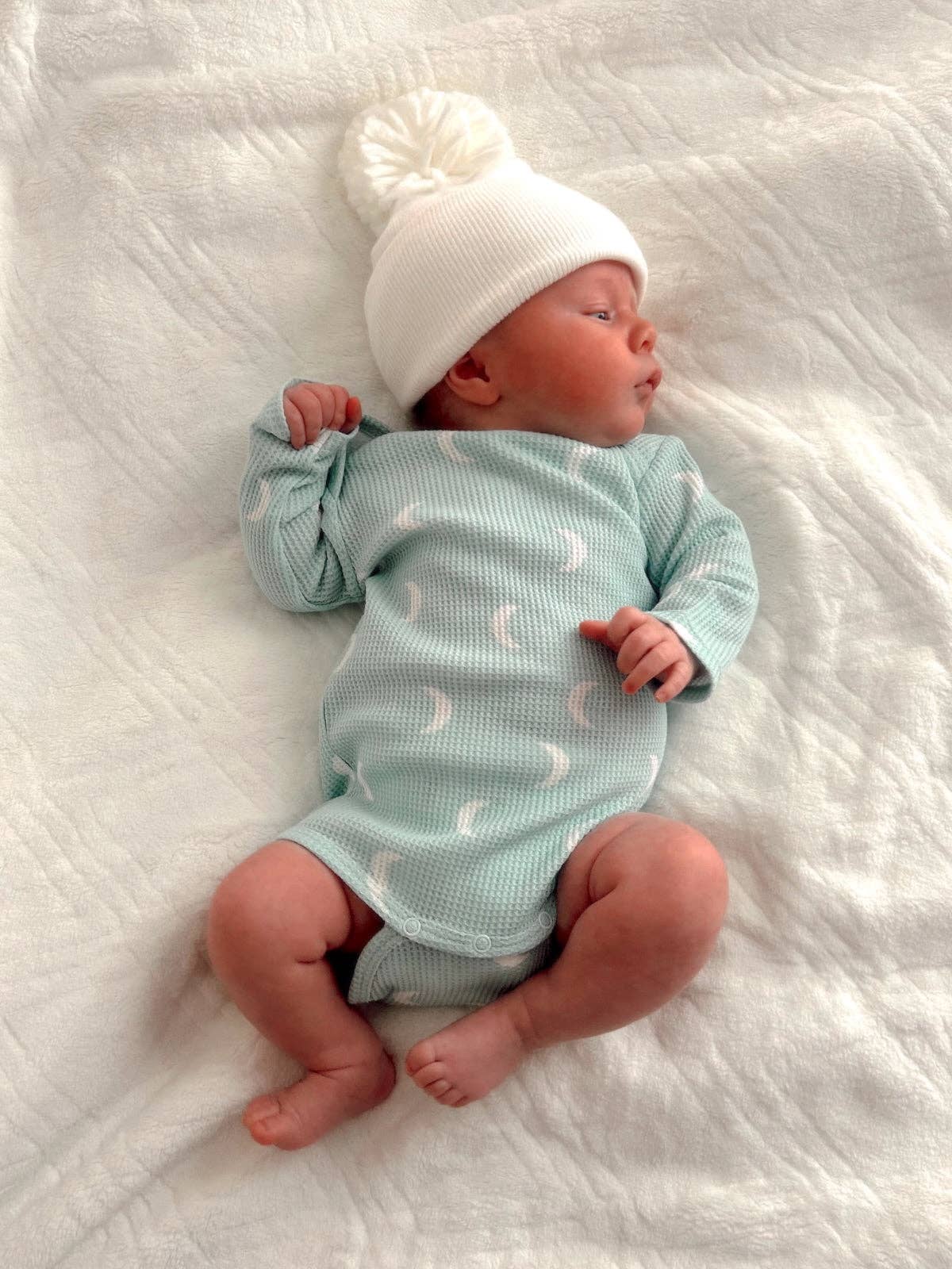 Newborn baby wearing a light blue onesie with white patterns and a white knit hat on a white textured blanket.