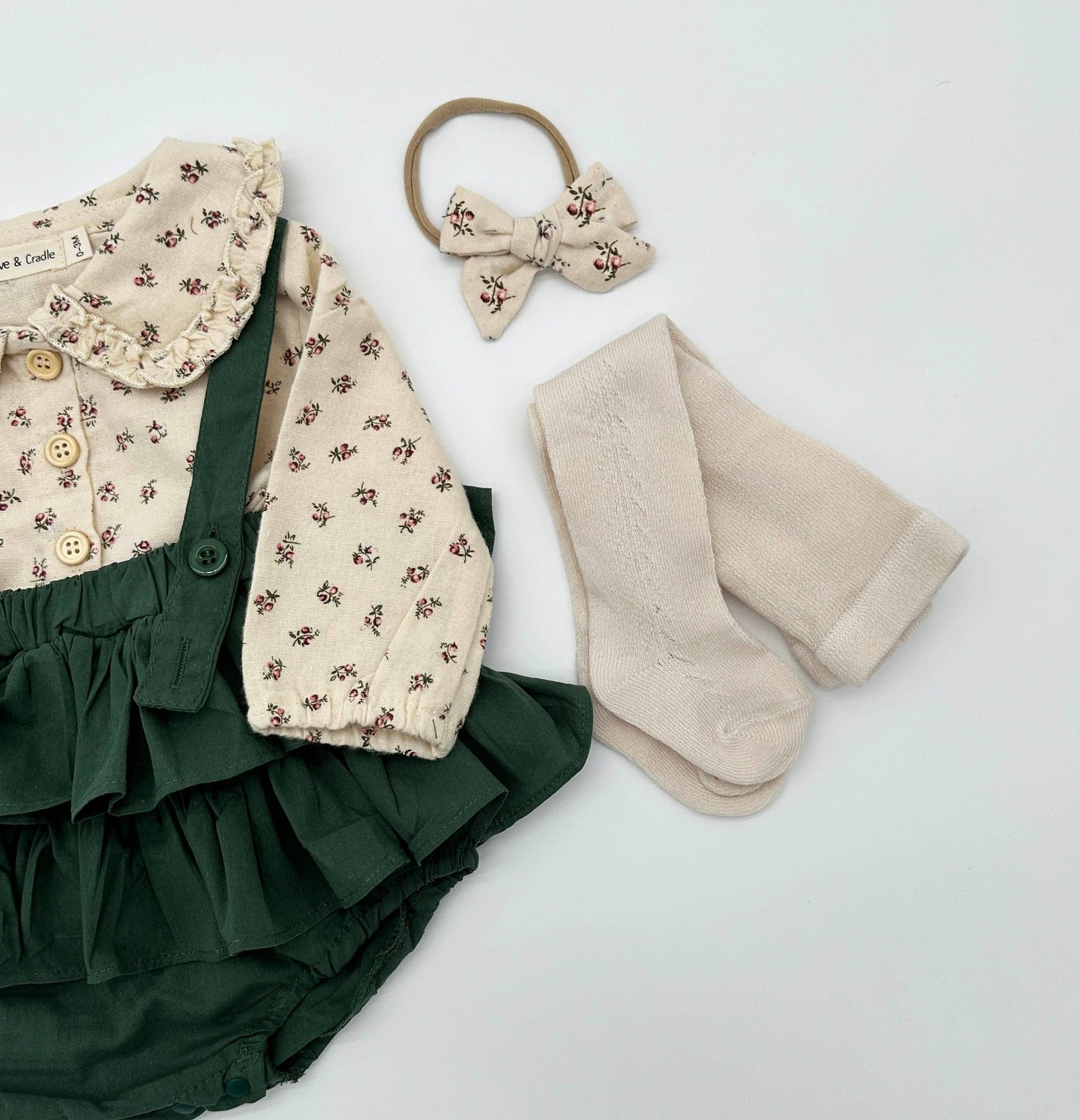 Baby outfit with green ruffled bloomers, floral top, beige socks, and hair accessories on a white background
