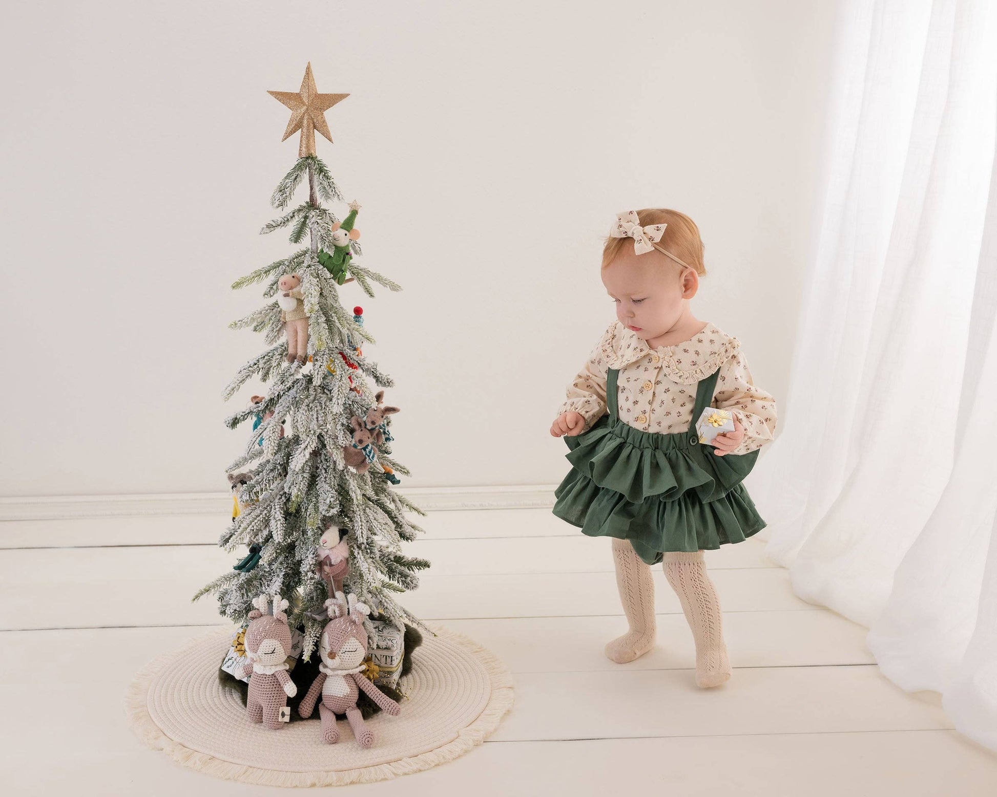 Child in a green dress standing next to a small decorated Christmas tree on a white background