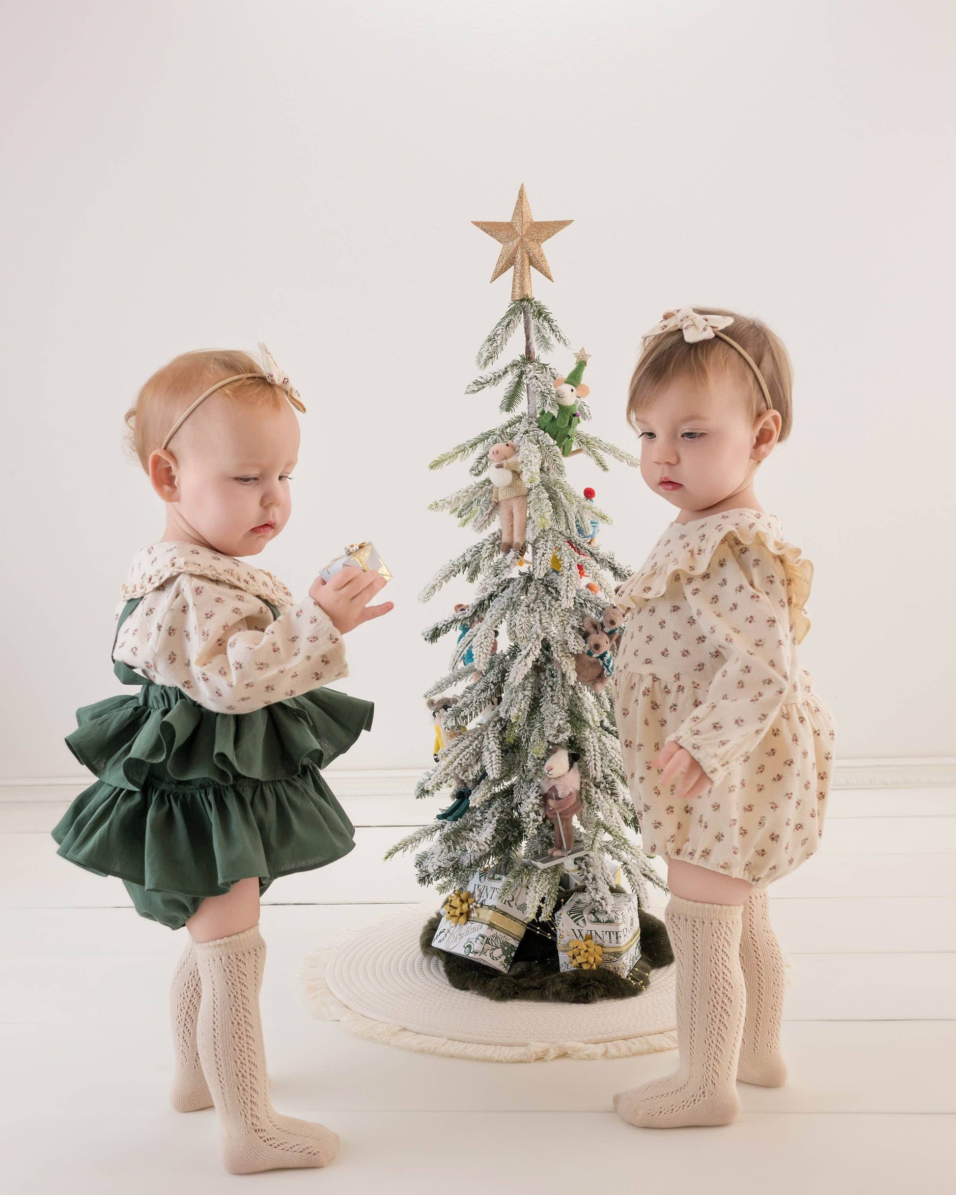 Two young children in matching outfits standing next to a small decorated Christmas tree.