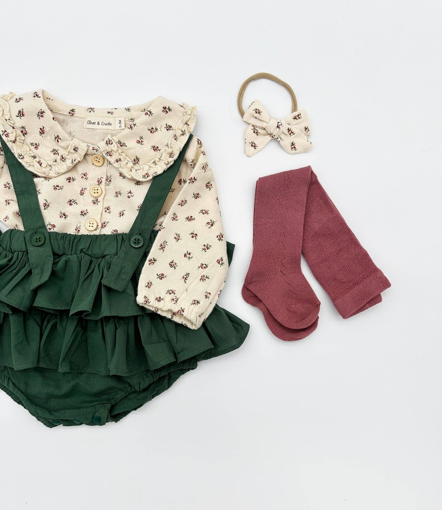Baby outfit set with green ruffled romper, floral shirt, socks, and hair tie on a light gray background