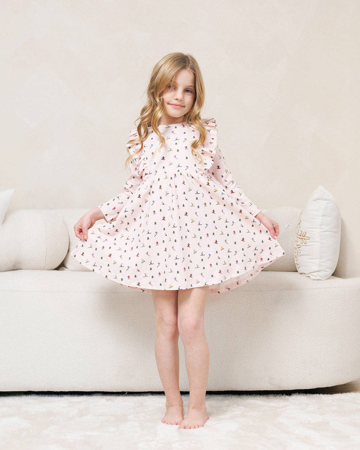 A young girl with blonde hair stands barefoot in front of a cream-colored sofa, wearing a long-sleeved, ruffled-neck babydoll dress in white with a small pink and brown floral pattern.