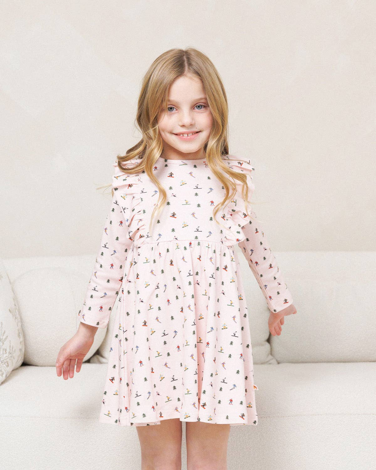 A young girl with blonde hair stands smiling in front of a light beige sofa, wearing a light pink long-sleeve dress with a small multicolor print and ruffled shoulders.