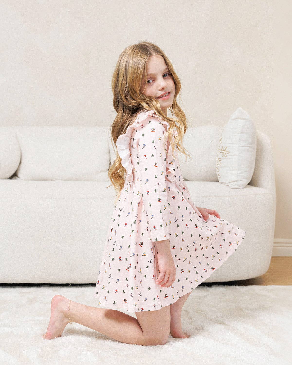 A young girl with long blonde hair kneels on a white rug in front of a cream-colored sofa, wearing a light pink long-sleeve dress with a small black and red pattern.
