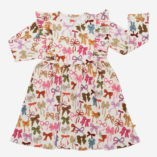 A long-sleeve baby dress with a ruffled neckline and a flared skirt, covered in an all-over print of colorful bows on a cream background.