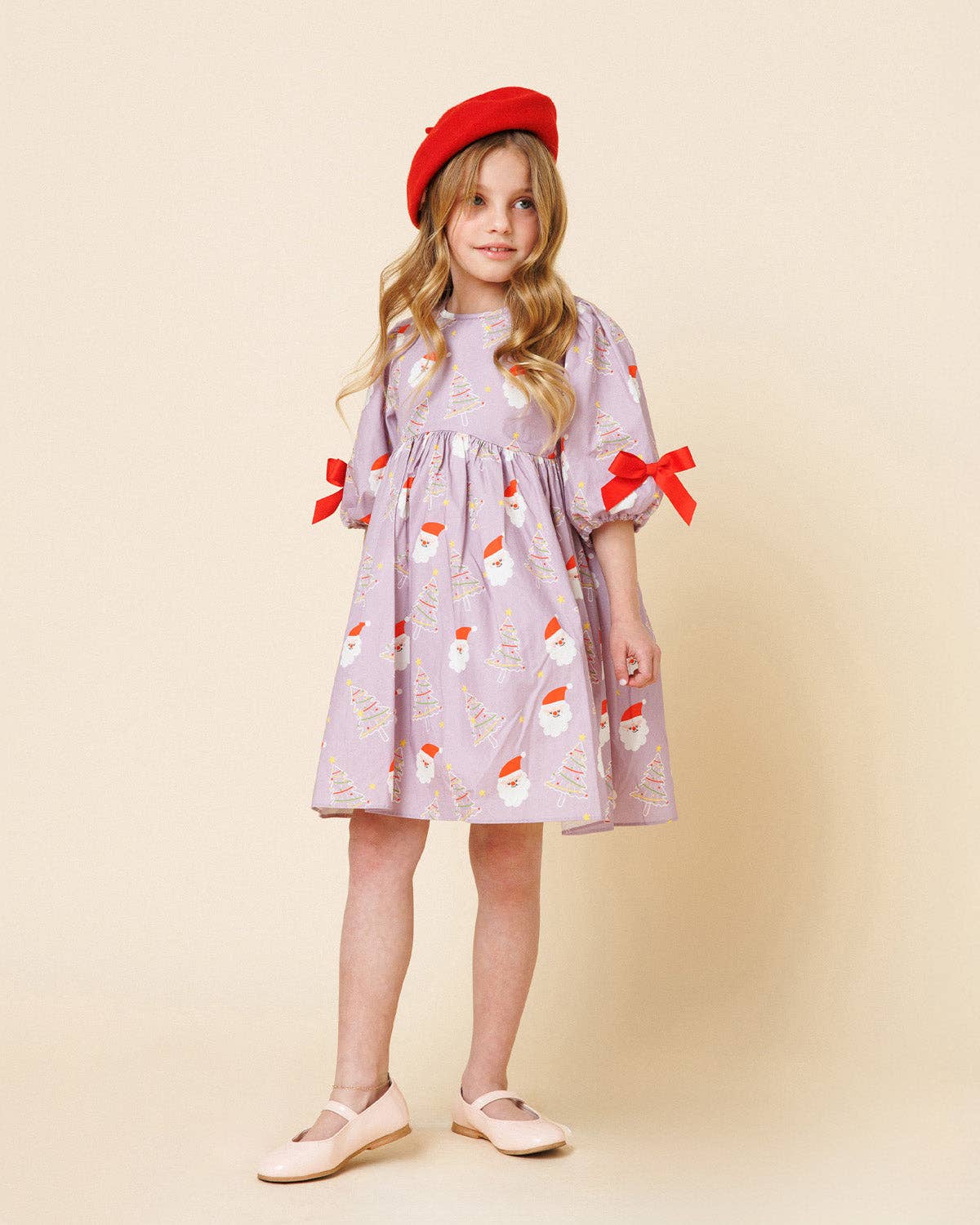 A young girl stands against a warm beige studio background wearing a light lavender dress with a Santa Claus print, red ribbon bows at the cuffs, and a red beret, paired with pale pink shoes.