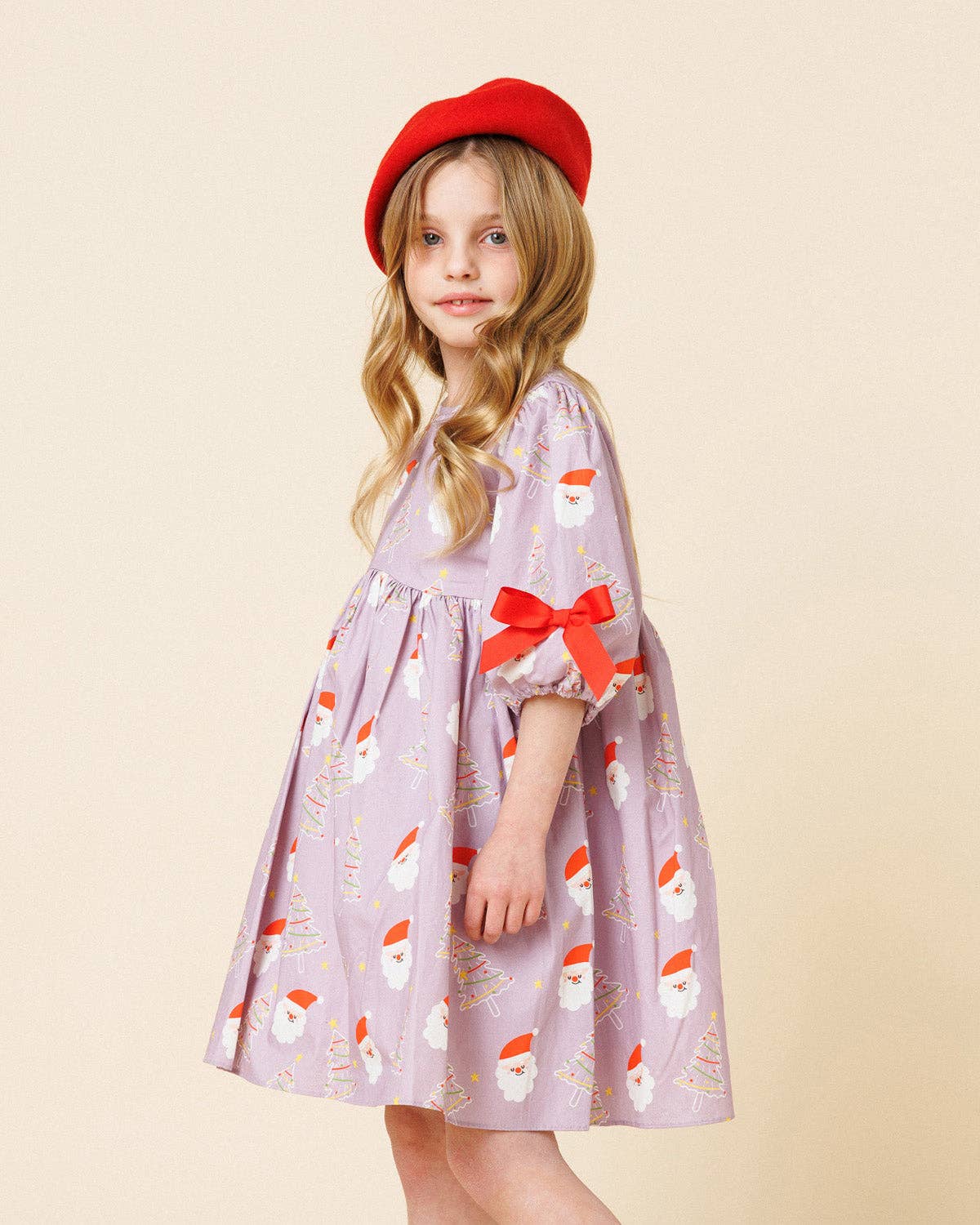 A young girl wearing a light purple Santa Claus patterned dress with a red bow on the sleeve and a red beret, standing against a solid beige background.
