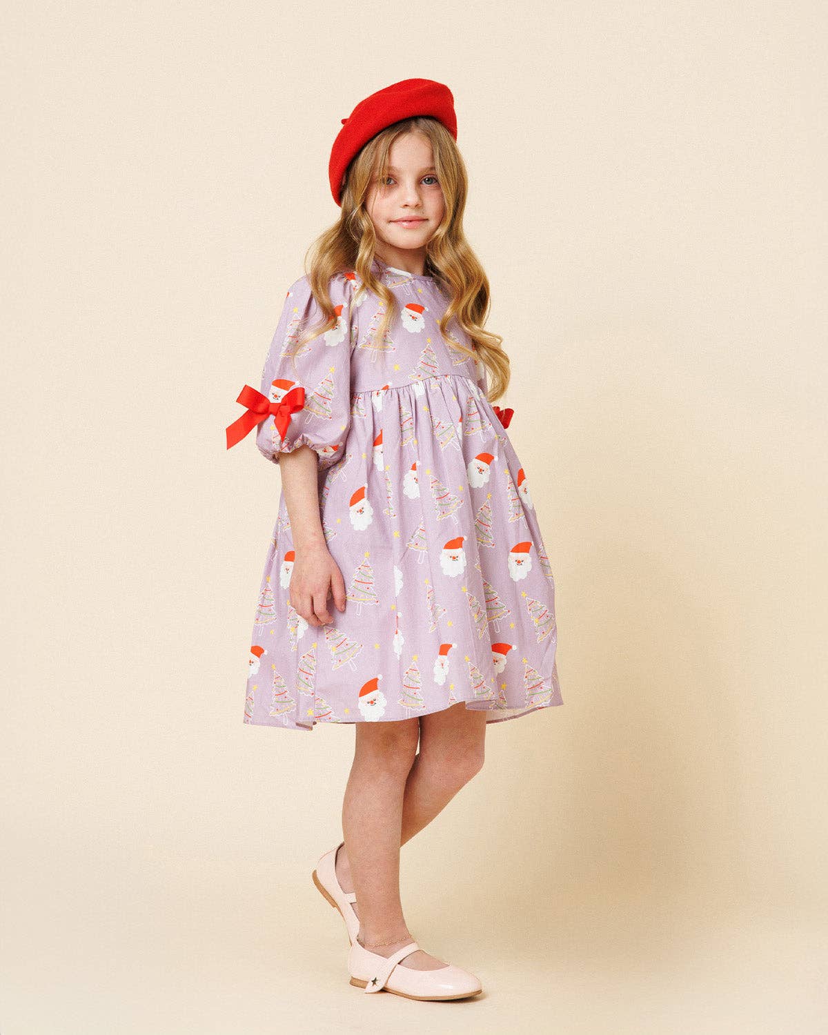 A young girl stands against a plain beige studio background wearing a lavender dress with a Santa Claus print, red bow details on the sleeves, a red beret, and pale pink shoes.