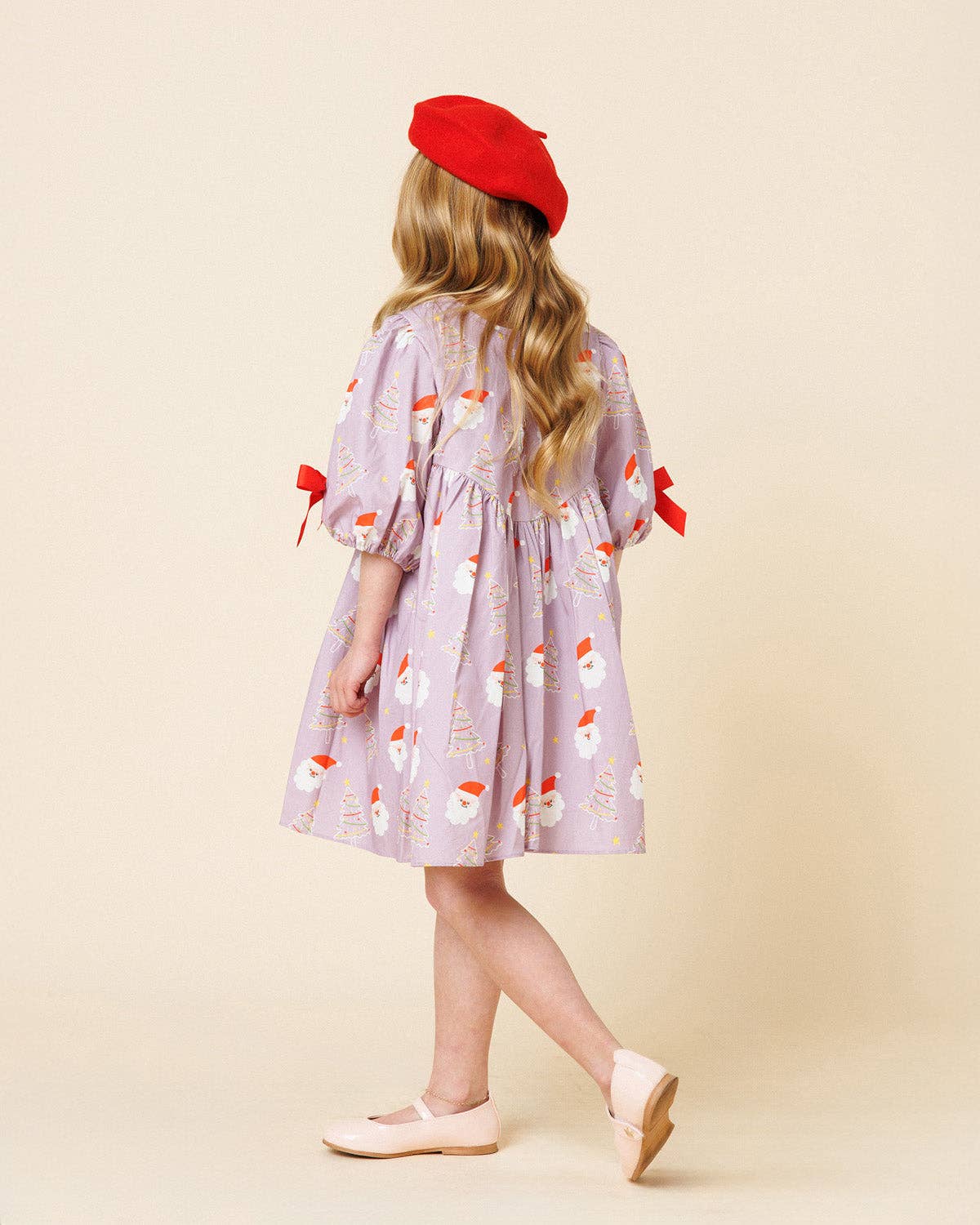 A young girl is shown from behind wearing a lavender puff-sleeve dress with a snowman pattern, a red beret, and light pink shoes, set against a solid beige background.