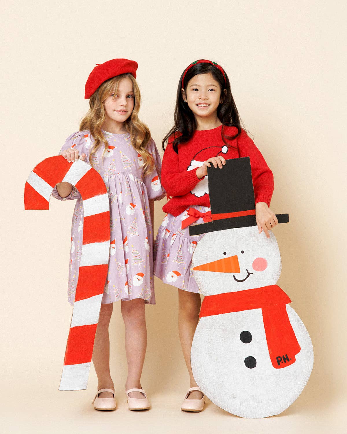 Two young girls modeling holiday costumes in a studio setting. The girl on the left wears a red beret and a light lavender dress with candy cane prints, holding a large red-and-white striped candy cane prop.