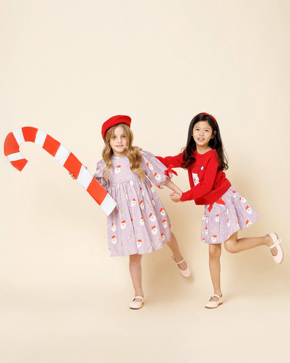 Two young girls modeling holiday-themed clothing against a solid beige studio background. The girl on the left wears a lavender Christmas print dress, a red beret, and holds a large red-and-white candy cane prop.