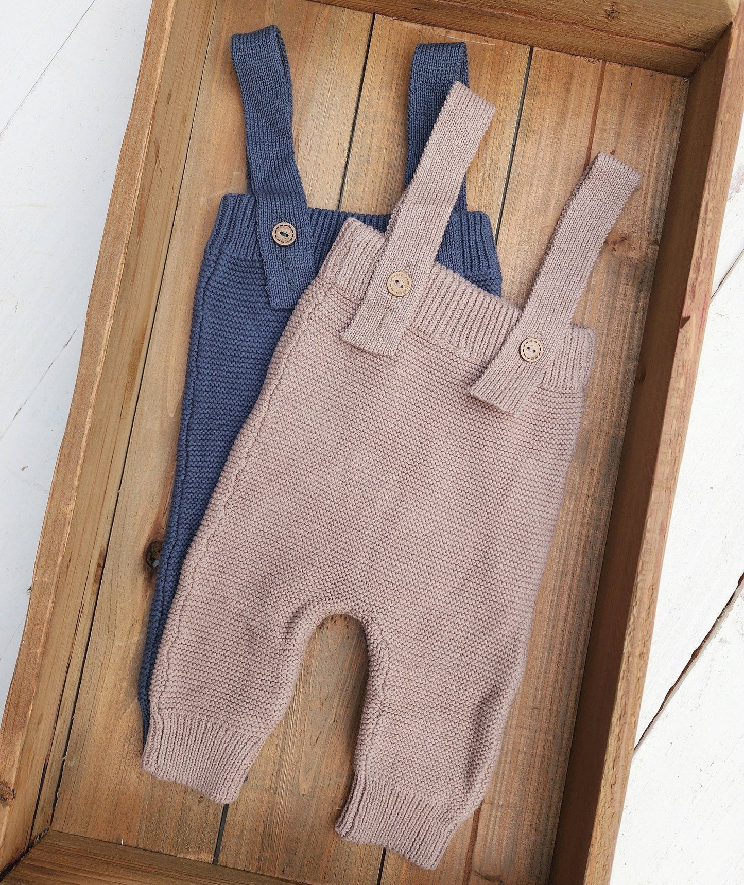 Two pairs of baby overalls, one blue and one brown, on a wooden surface.