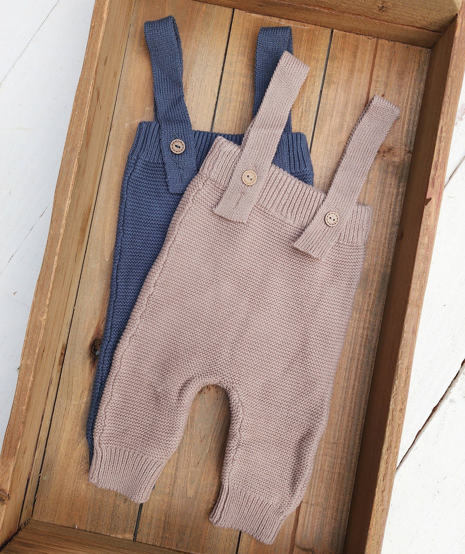 Two pairs of baby overalls, one blue and one brown, on a wooden surface.