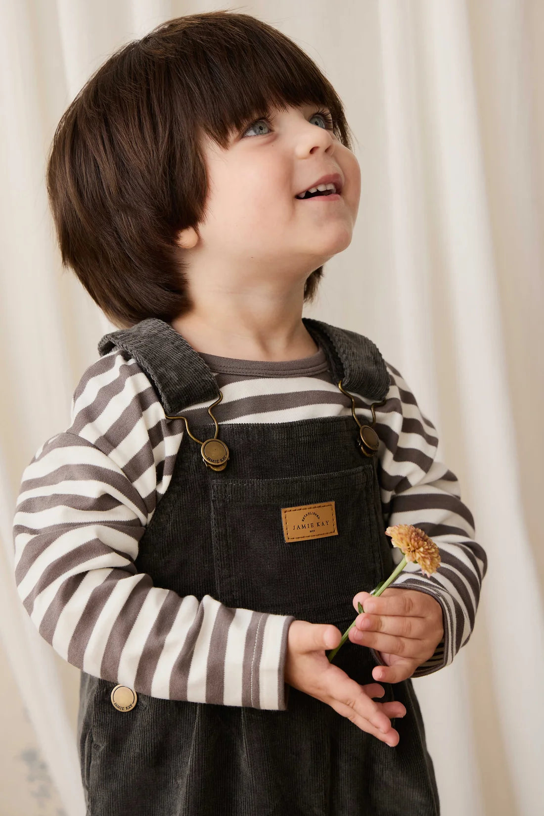 Child wearing a striped shirt and denim overalls holding a flower against a white curtain background