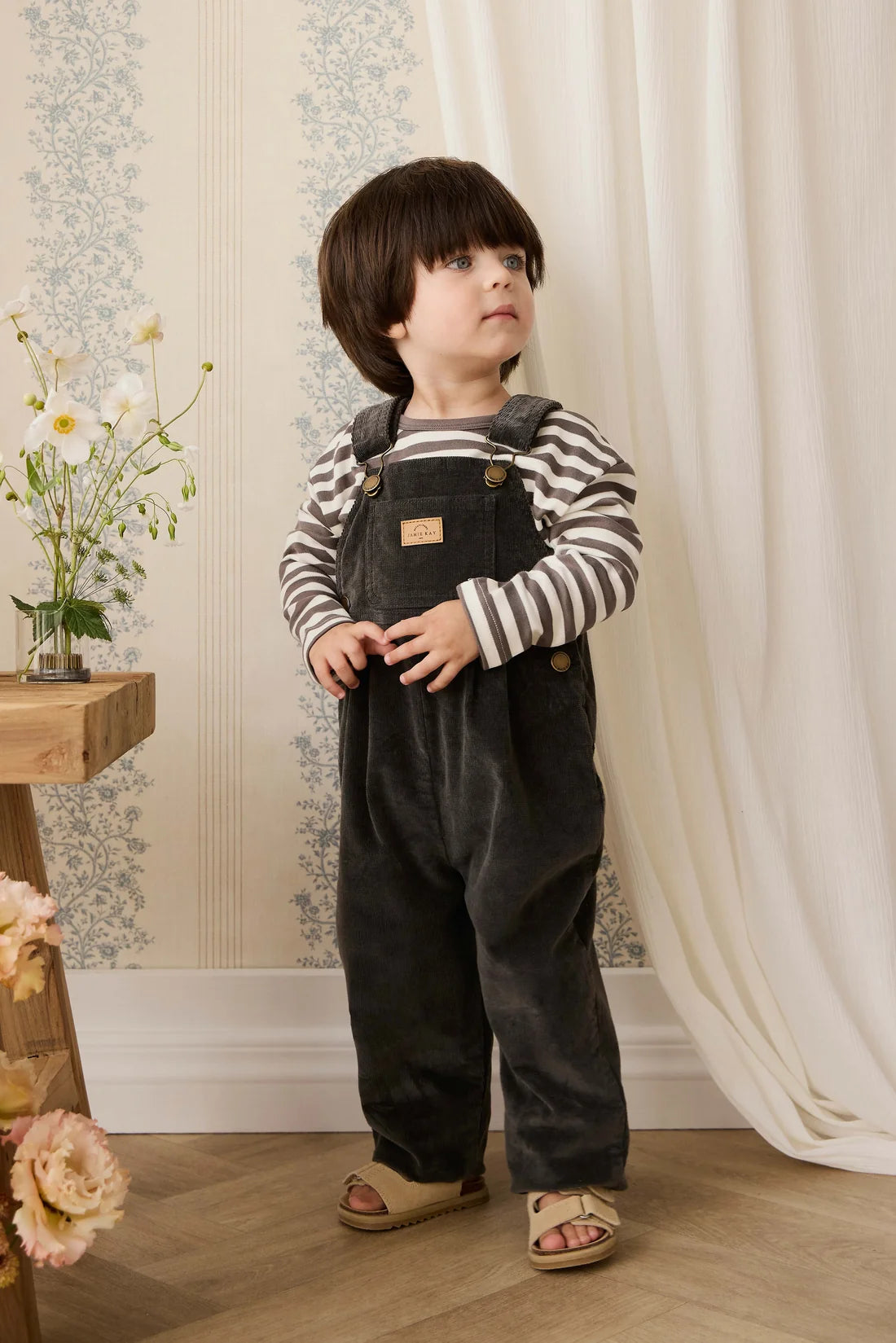 Child wearing black overalls and striped shirt standing in a room with floral decorations.
