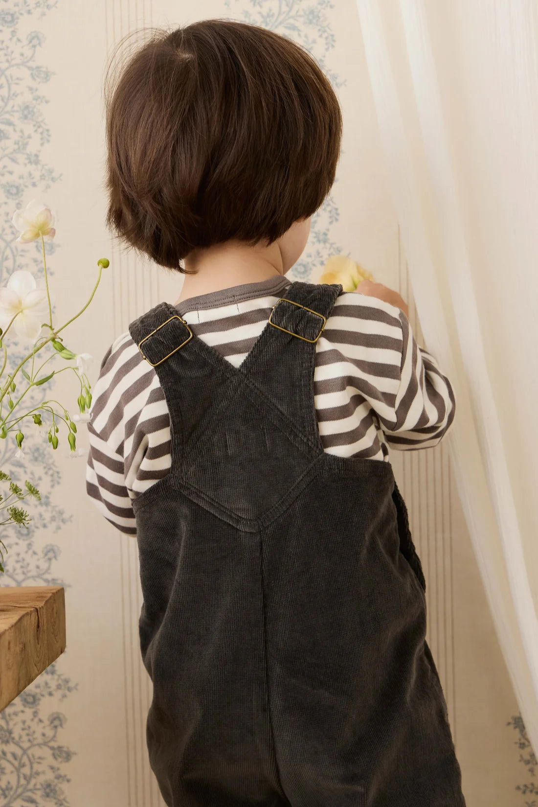 Child wearing black overalls and a striped shirt, standing in front of a floral-patterned wall.