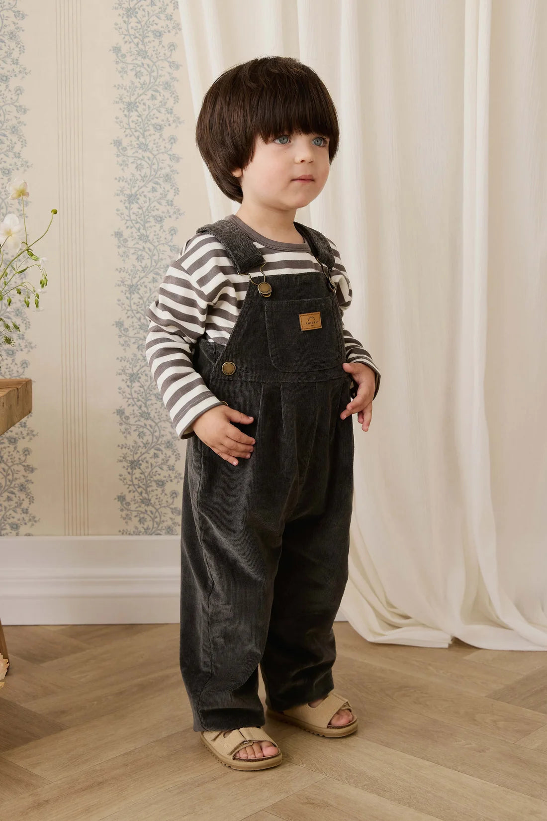 A toddler standing indoors wearing dark charcoal corduroy overalls over a gray and white striped long-sleeve shirt, paired with beige sandals. The background features cream-colored curtains and patterned wallpaper.