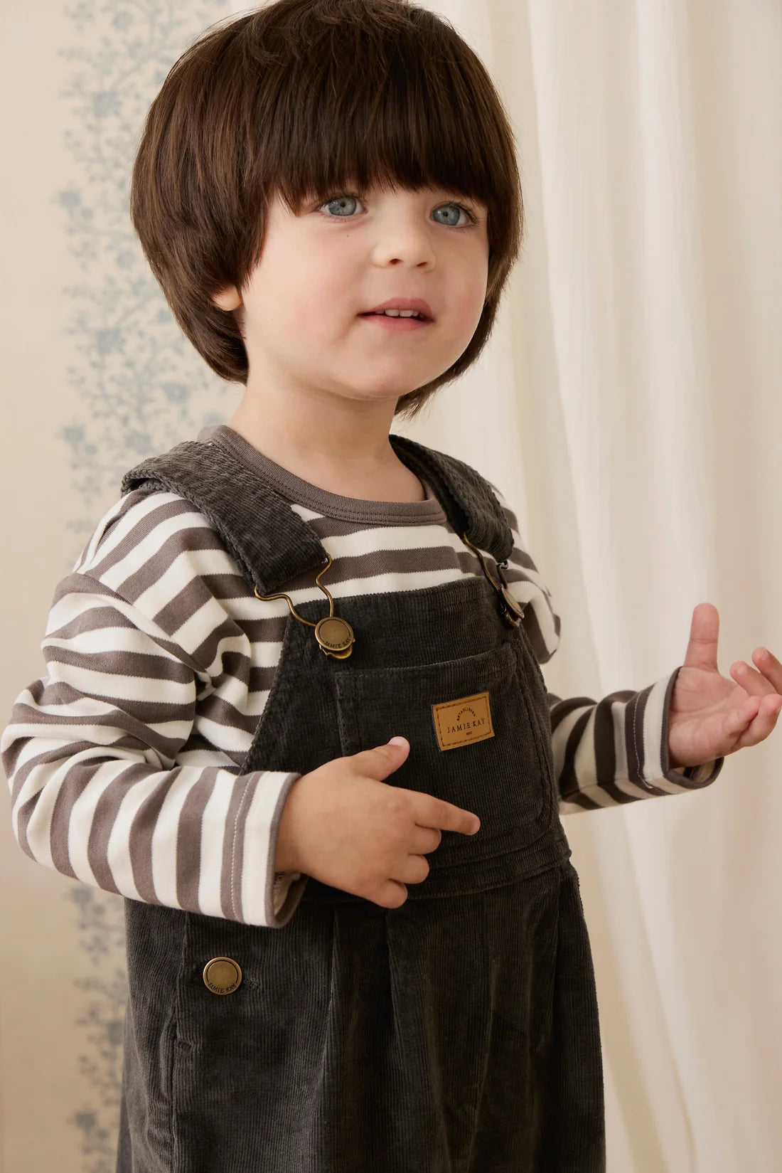 A toddler with dark brown hair and blue eyes wearing dark gray corduroy overalls over a brown and white striped long-sleeve shirt, standing indoors against a cream curtain background.