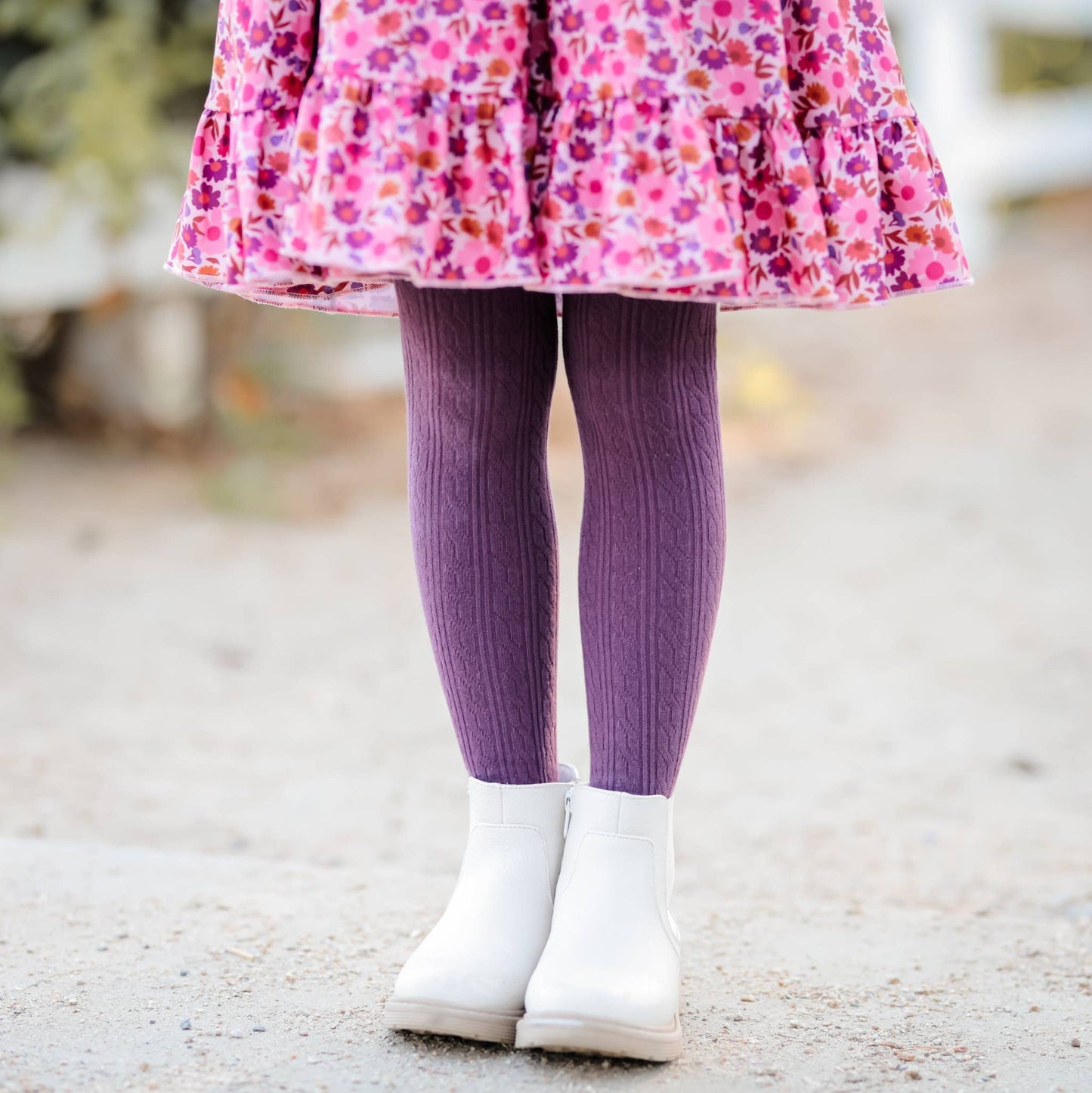 Child wearing a pink floral dress, purple tights, and white boots on a blurred outdoor background