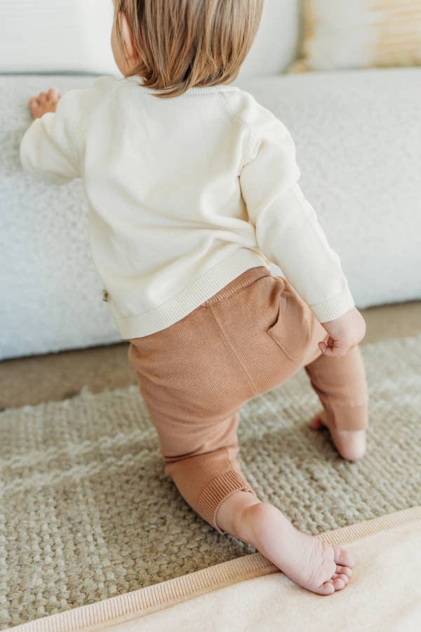 Child wearing a cream sweater and brown pants standing on a carpeted floor.