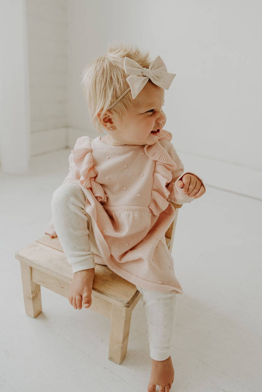 Baby in a pink sweater with ruffles sitting on a wooden stool against a white background