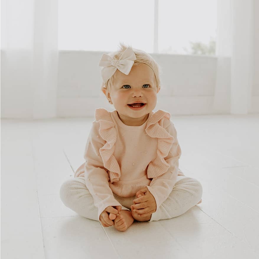 Baby sitting on a white floor wearing a pink ruffled outfit with a white bow.