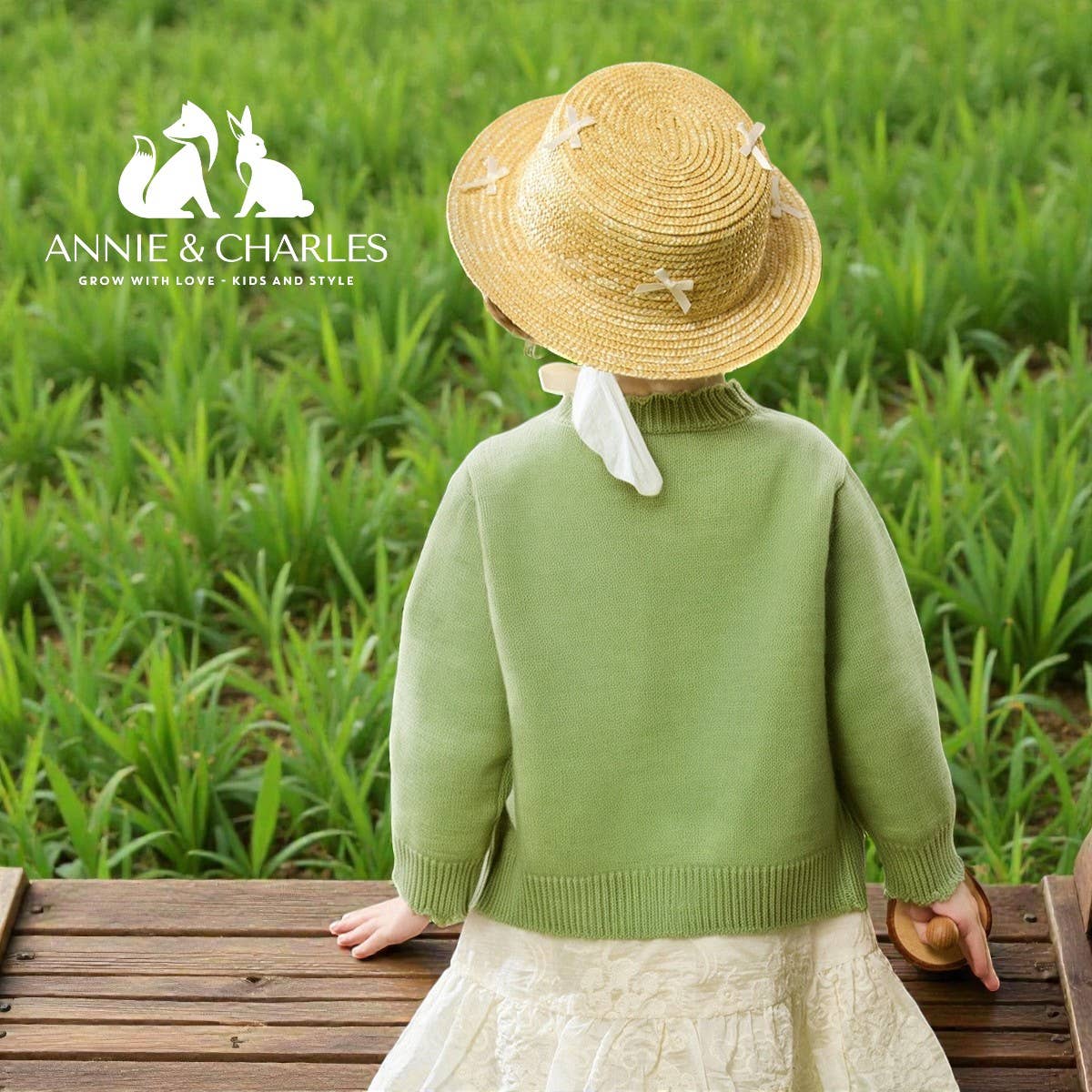 Child wearing a straw hat and green sweater in a grassy field with Annie & Charles logo.