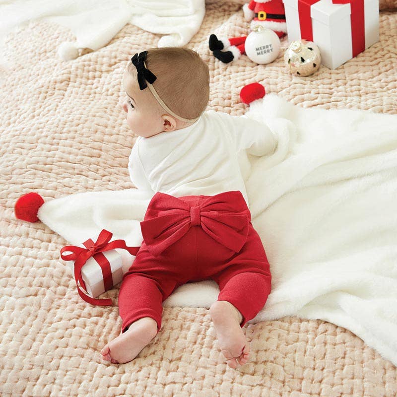 Baby in a white outfit with a large red bow on a beige blanket