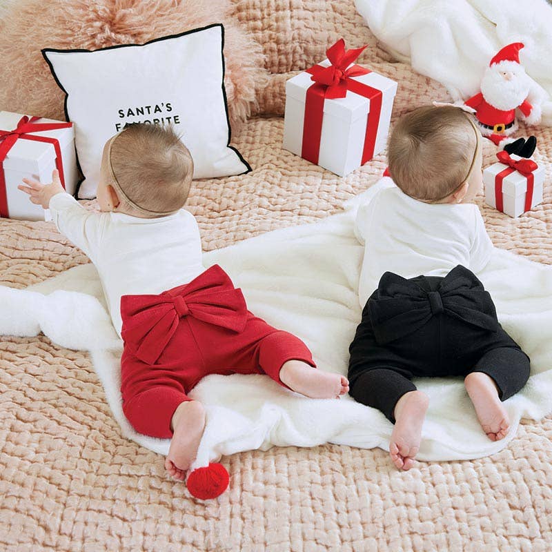 Two babies in Christmas-themed outfits lying on a blanket with presents and a Santa pillow in the background.