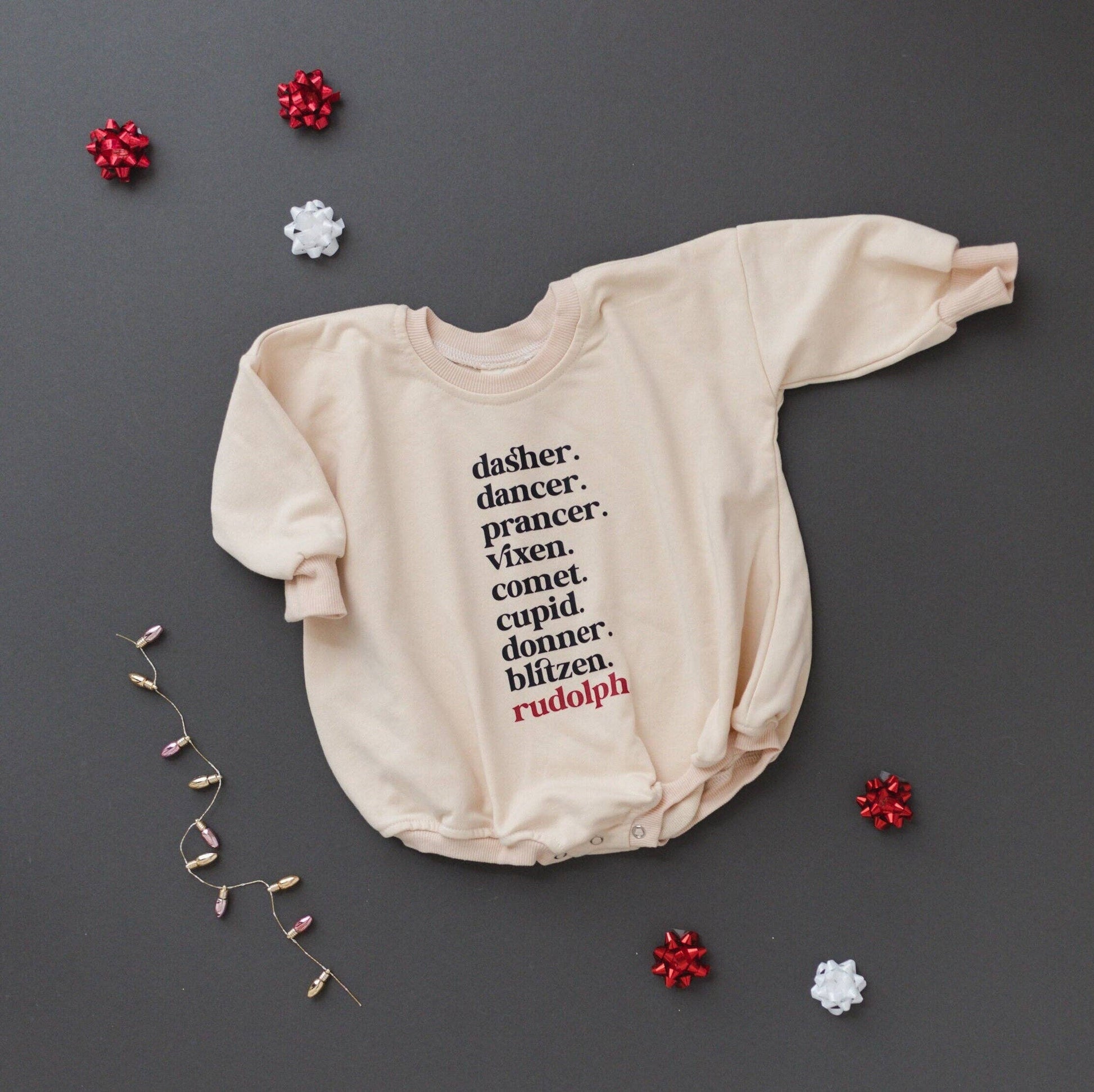 Beige baby onesie with Christmas-themed text on a dark gray background with decorative elements.