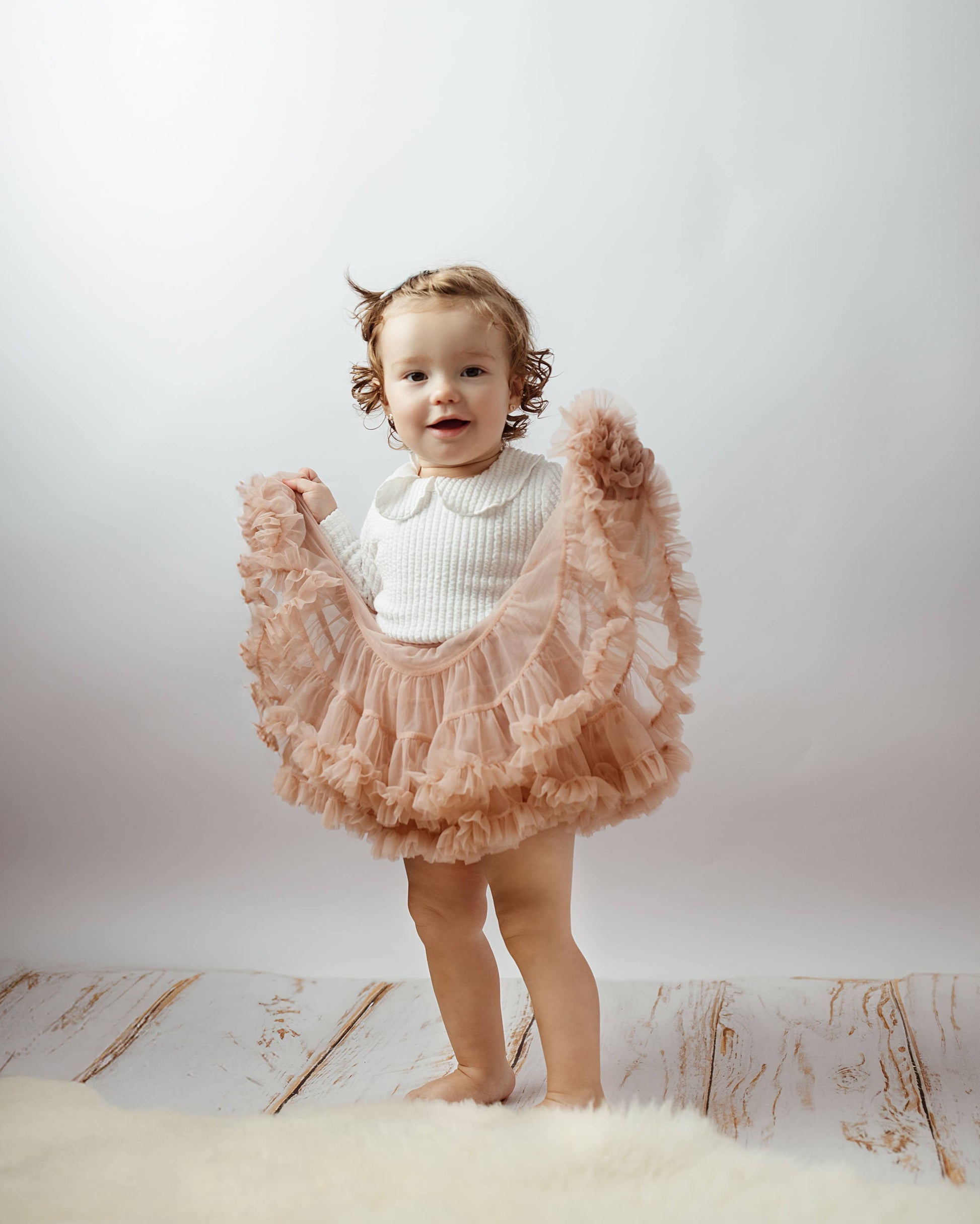 Child wearing a white top and pink ruffled skirt on a light wooden floor with a white background