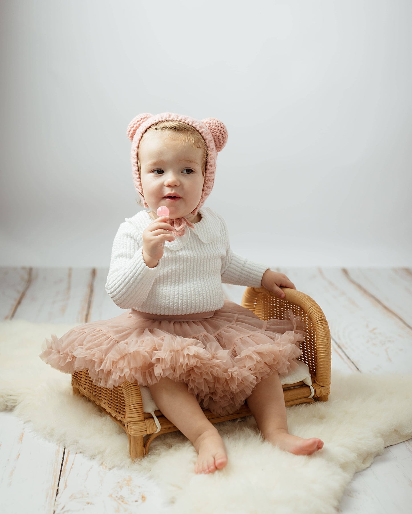 Baby in a pink tutu and bear hat sitting on a wicker stool against a white background