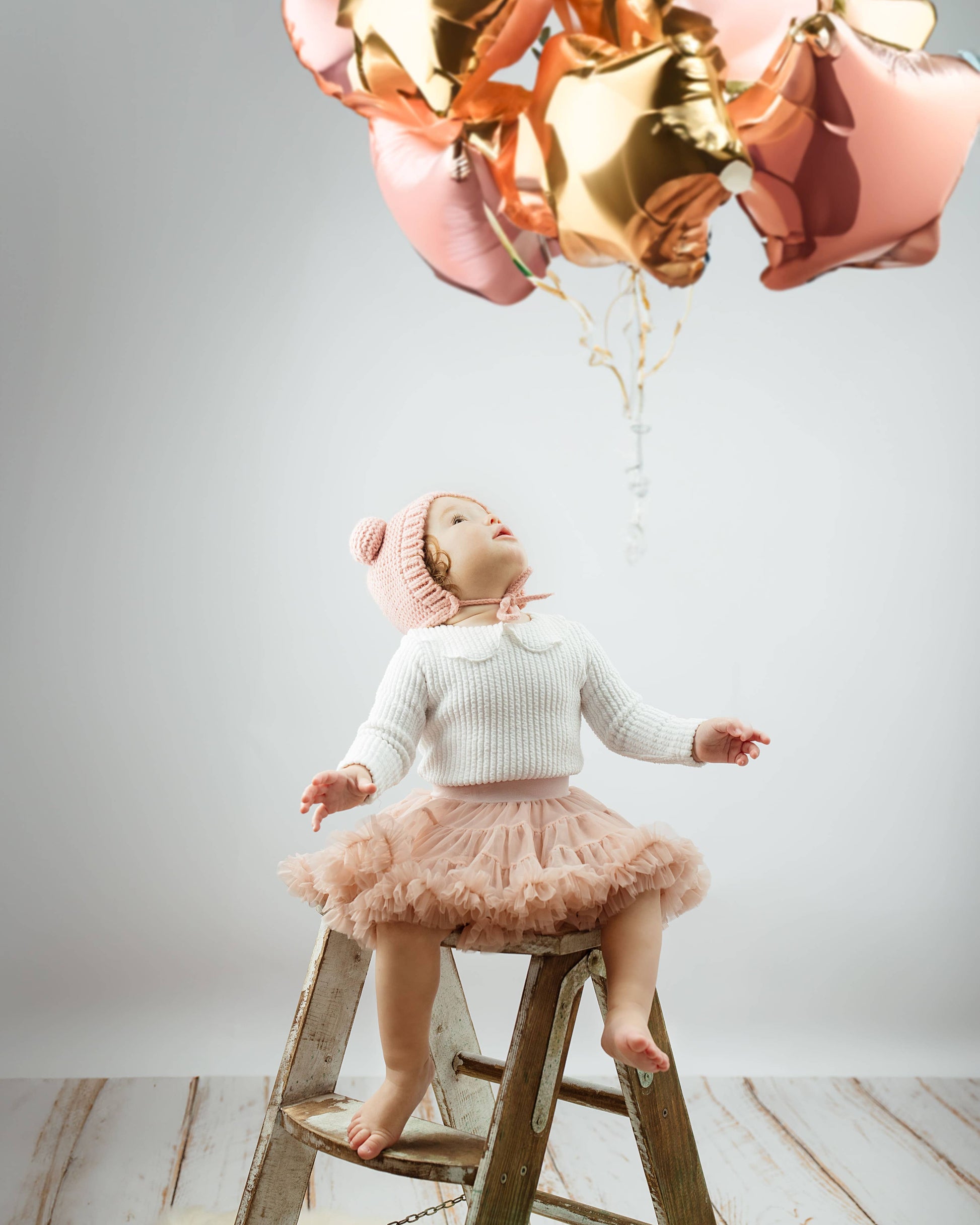 Child on a ladder with pink and gold balloons against a white wall