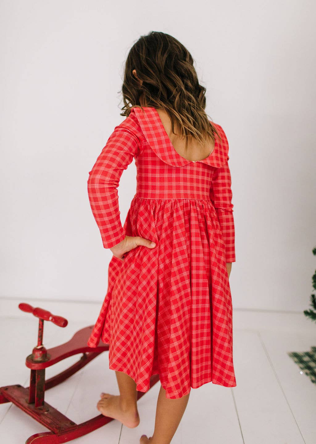 Person wearing a red checkered dress standing next to a red rocking horse on a white floor.