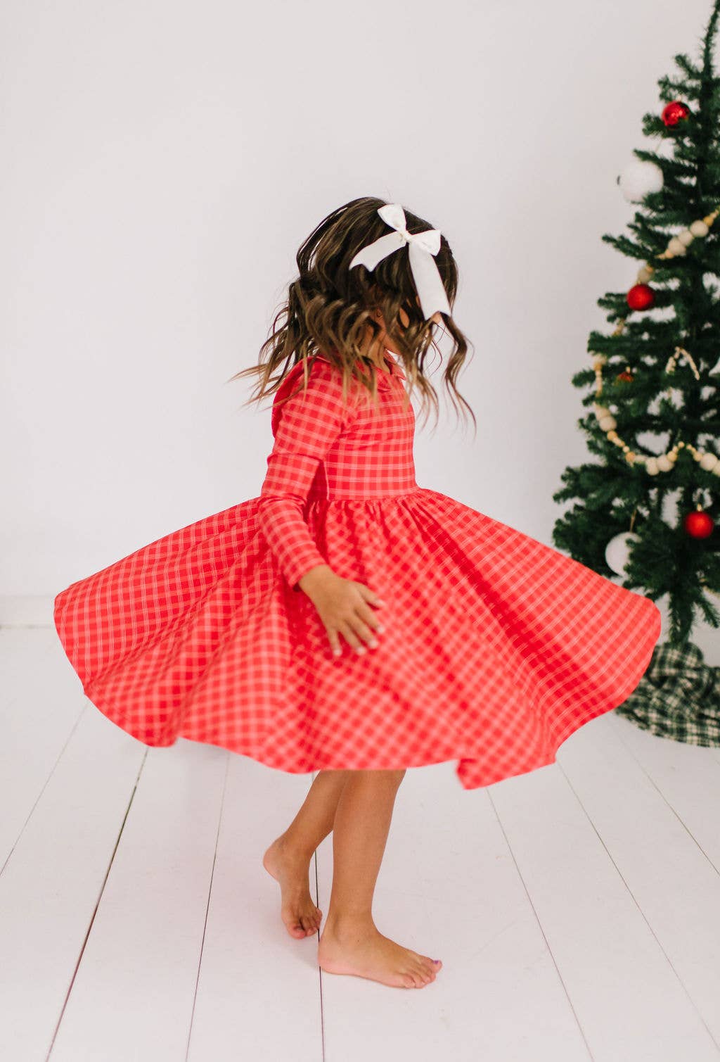 Child in a red checkered dress standing in front of a Christmas tree.