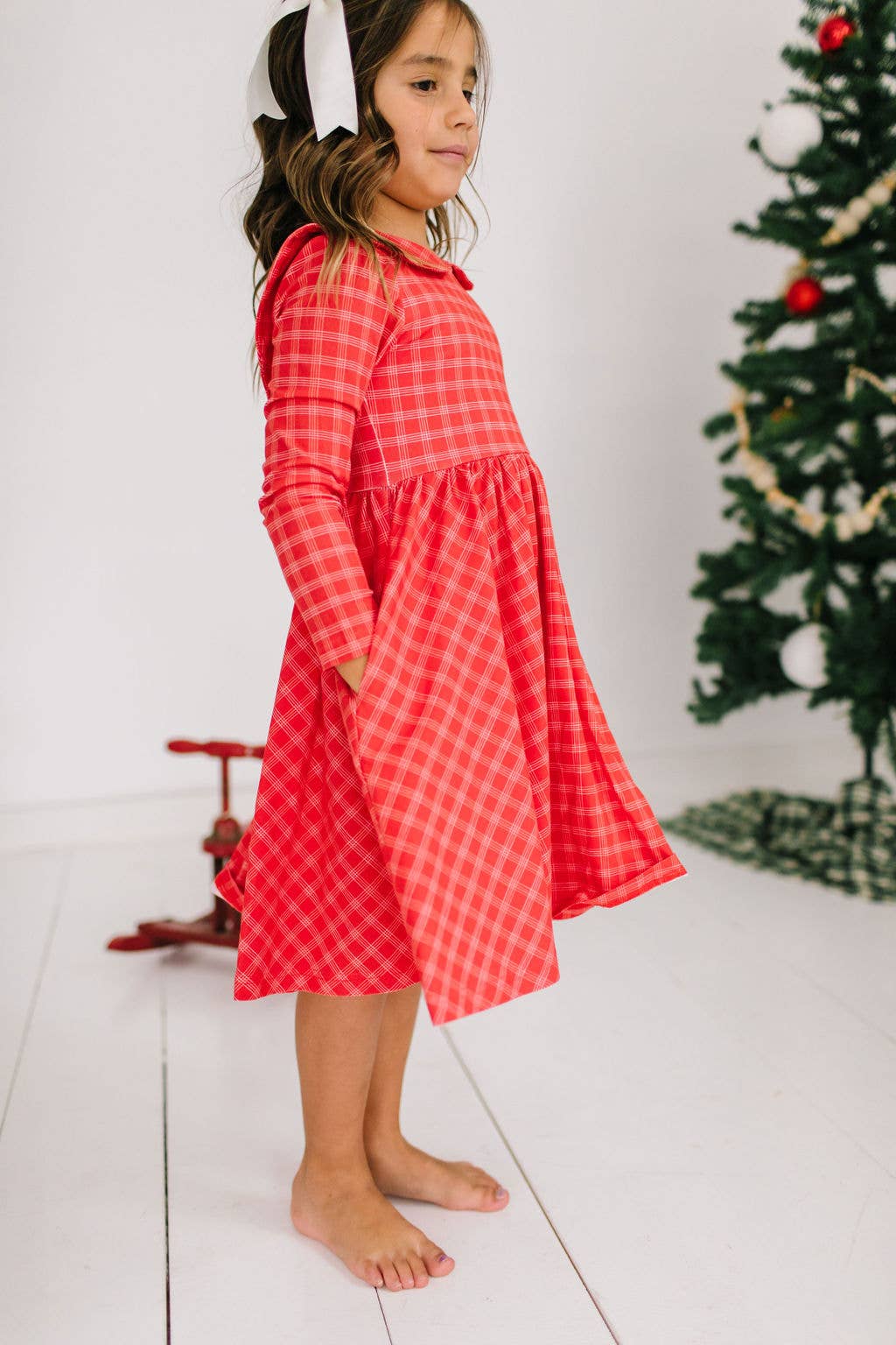 Young girl wearing a red checkered dress standing in front of a Christmas tree.